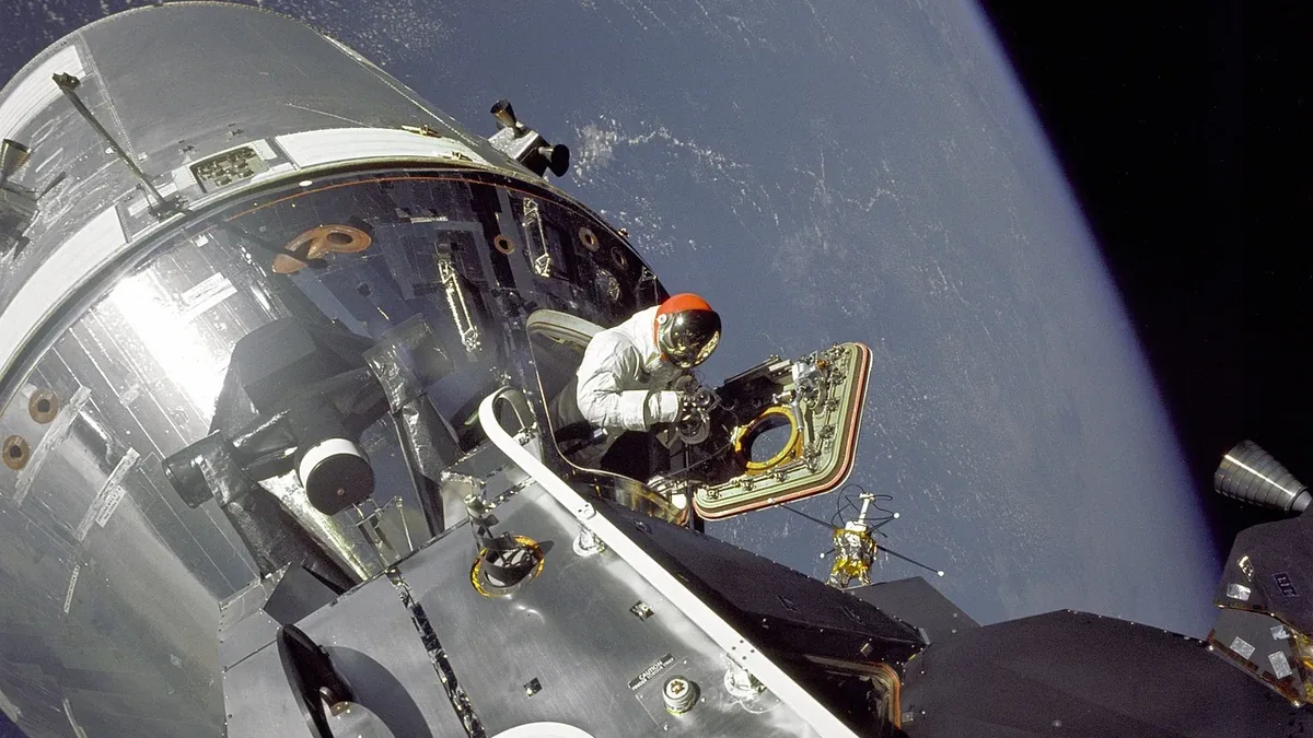 An astronaut is seen working on the exterior of a spacecraft in space, with the Earth visible in the background.