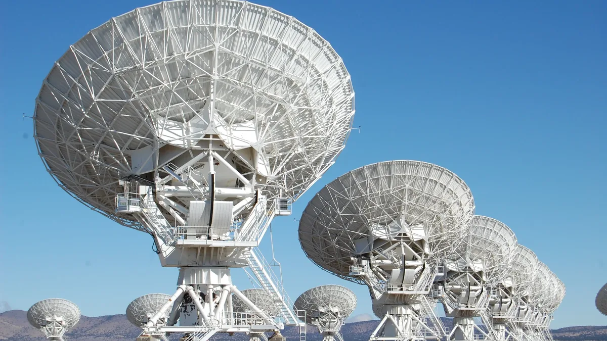 A group of large satellite dishes, likely used for radio astronomy or communication, situated in a desert-like setting with mountains visibl
