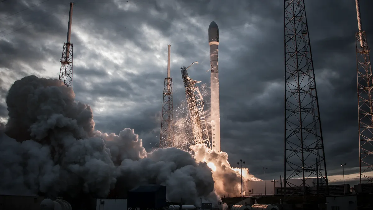 A rocket launch, with the rocket visible in the center, emitting a large plume of smoke and flames as it lifts off from a launchpad surround