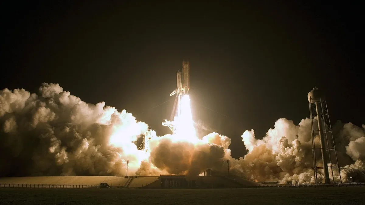 A rocket launch, with the rocket visible in the center, emitting a large plume of smoke and flames as it takes off from a launchpad.