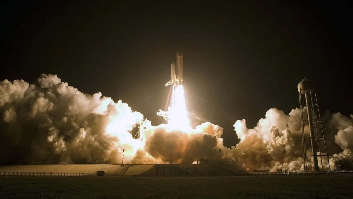 A rocket launch, with the rocket visible in the center, emitting a large plume of smoke and flames as it takes off from a launchpad.