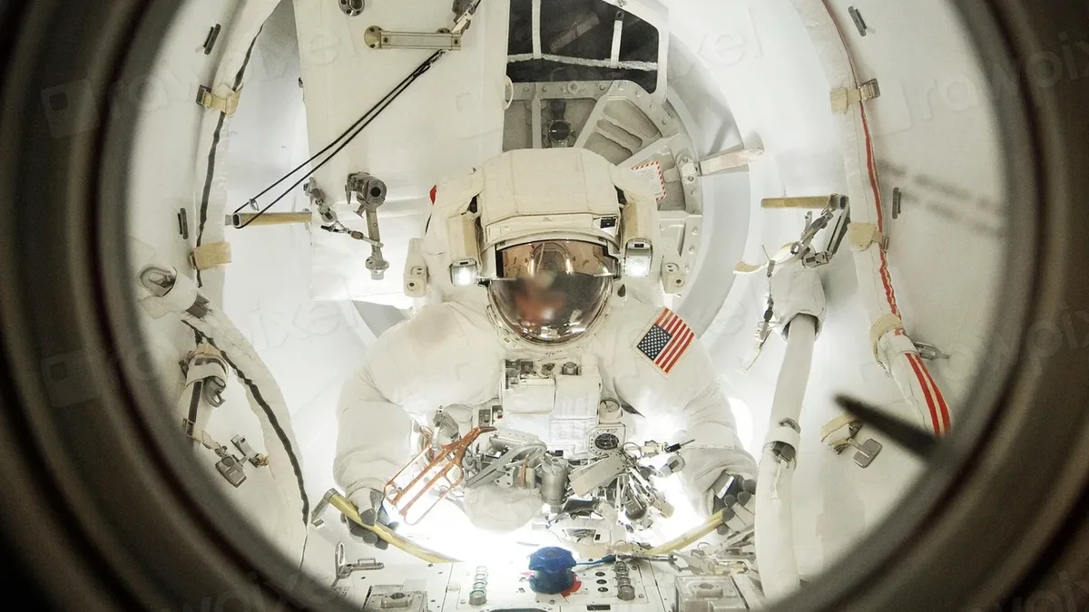 An astronaut in a white spacesuit, viewed through a round window or porthole, with the astronaut's helmet and visor reflecting light.